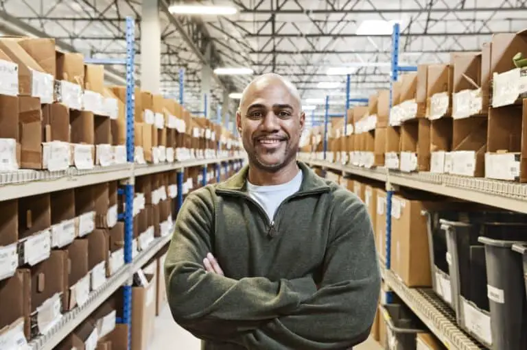 A black warehouse worker standing near boxed products in a distribution warehouse.